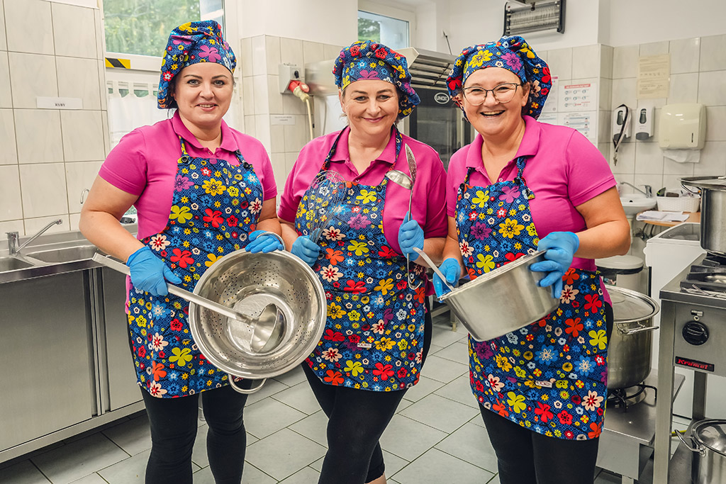 Trzy uśmiechnięte kobiety w kuchni gastronomicznej, ubrane w różowe koszulki i kolorowe fartuchy w kwiaty, trzymają garnki i przybory kuchenne, przygotowując posiłek.